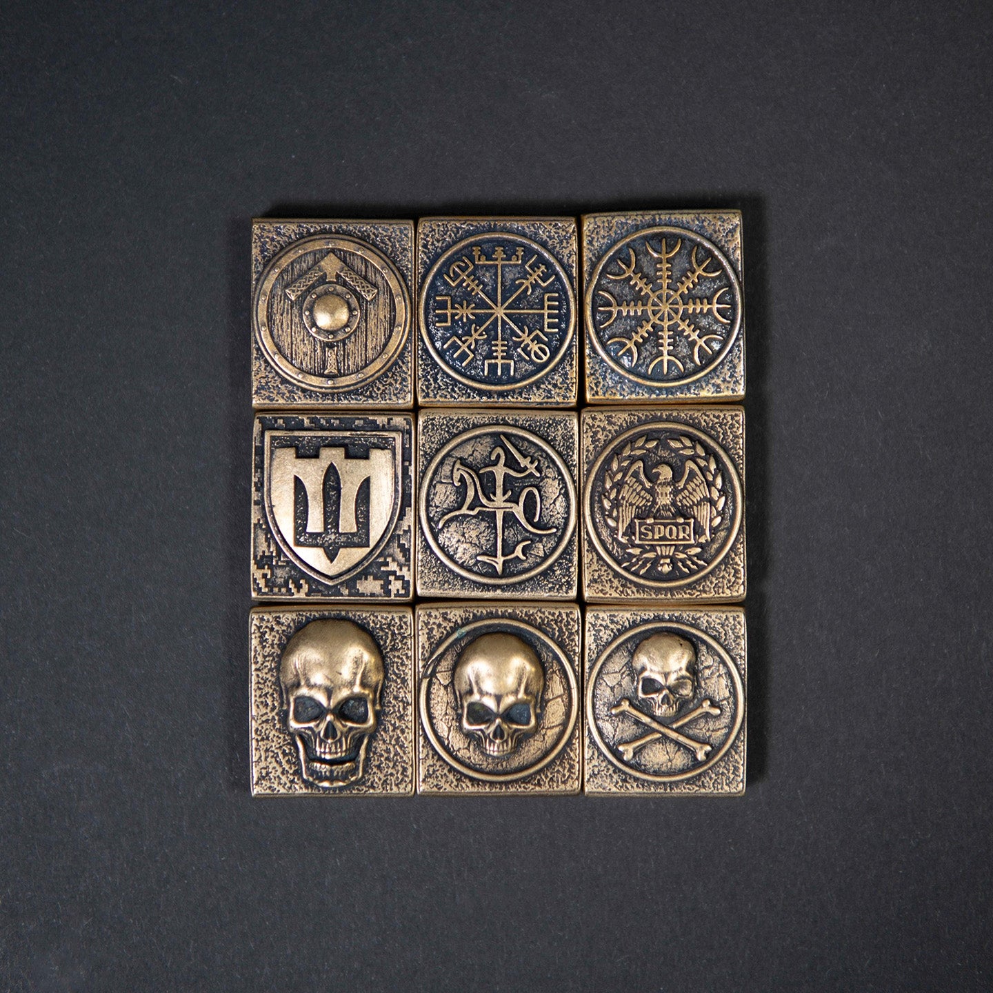 Set of solid brass MOLLE patches with antique finish, featuring skulls, Viking Vegvisir, shield, and crossbones designs displayed on a dark background.