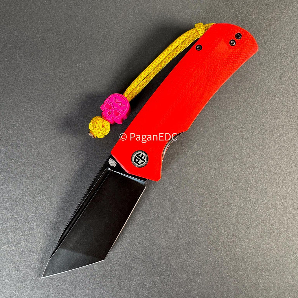 Folding knife "Beluga K110"  Red-handled with black tanto-style blade, yellow paracord lanyard, and pink PaganEDC skull bead on a gray background.