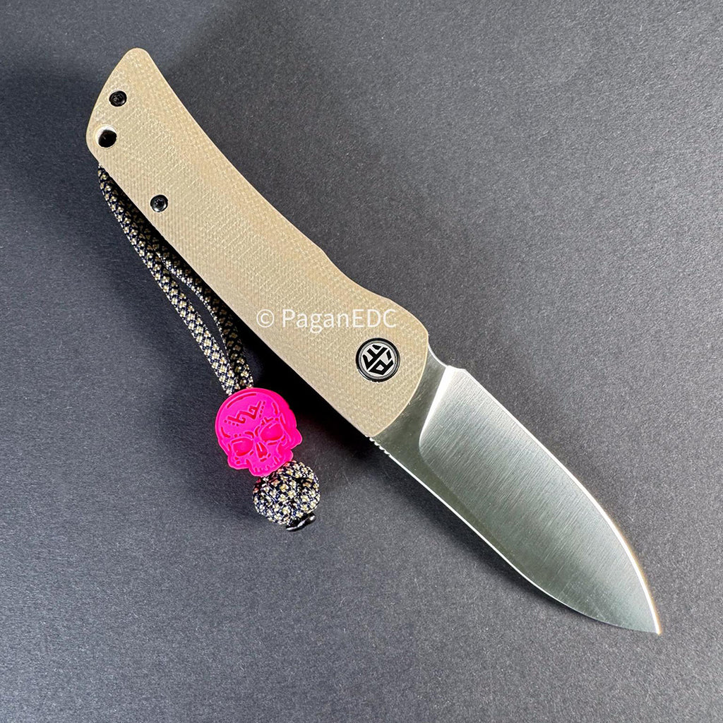 Tan-handled folding knife with a satin blade lying on a dark background, featuring a black-and-tan braided lanyard with a bright pink skull bead attached to the handle.