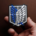 PVC patch featuring the blue and white winged emblem from Attack on Titan, held between fingers against a brown background