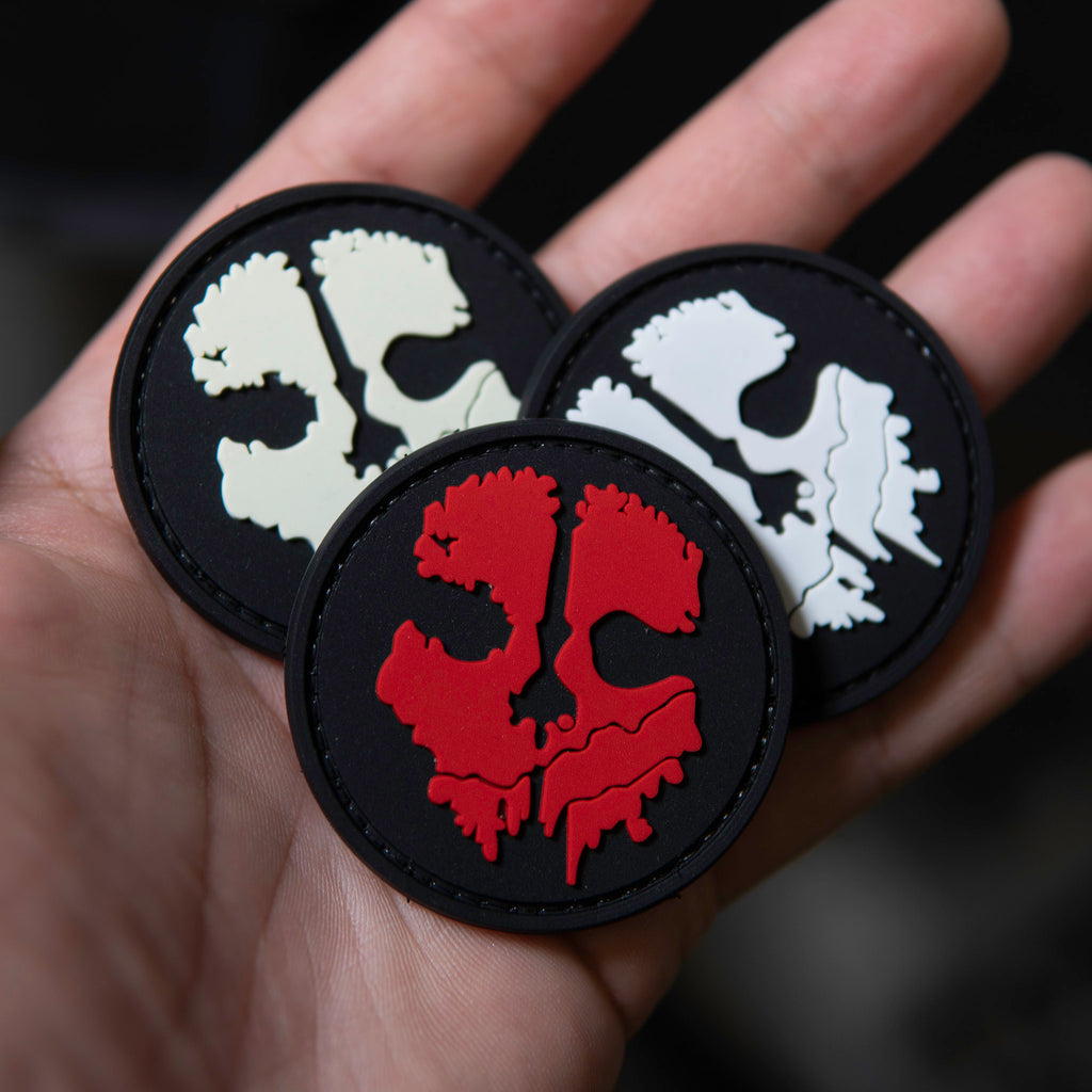 Hand holding three round PVC COD skull patches in white, red, and glow-in-the-dark colors on a black background.