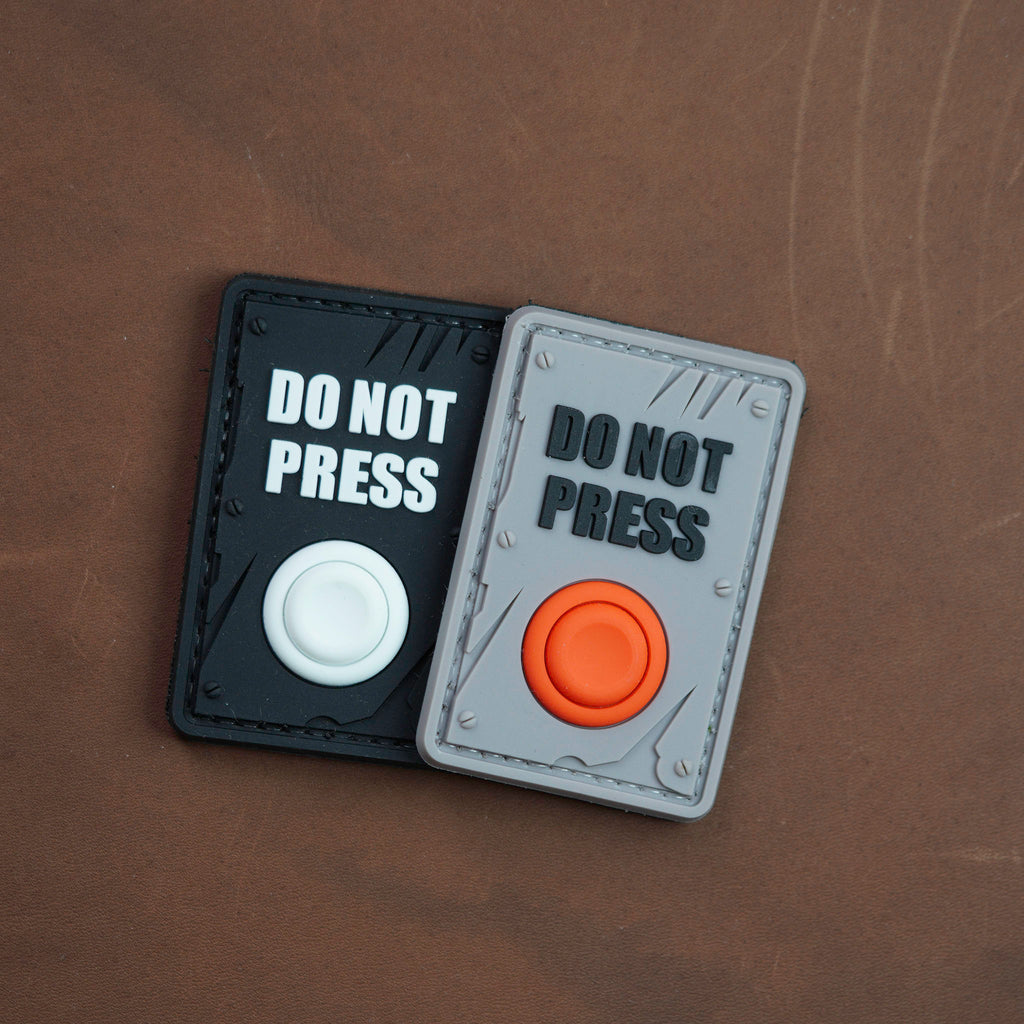 Two PVC patches labeled ‘DO NOT PRESS,’ one black with a white button and one grey with an orange button, placed on a brown surface.