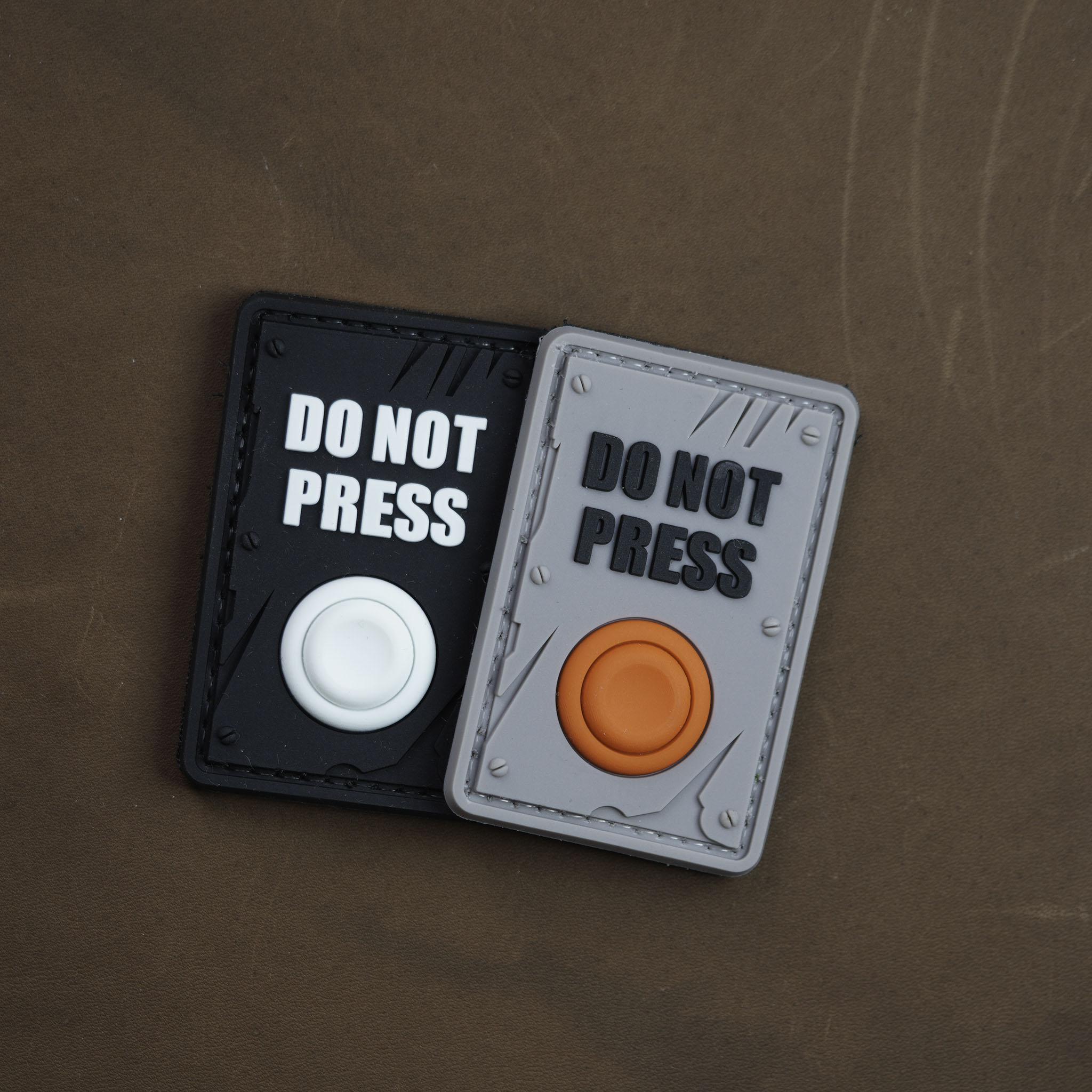 Two PVC patches labeled ‘DO NOT PRESS,’ one black with a white button and one grey with an orange button, placed on a brown surface.