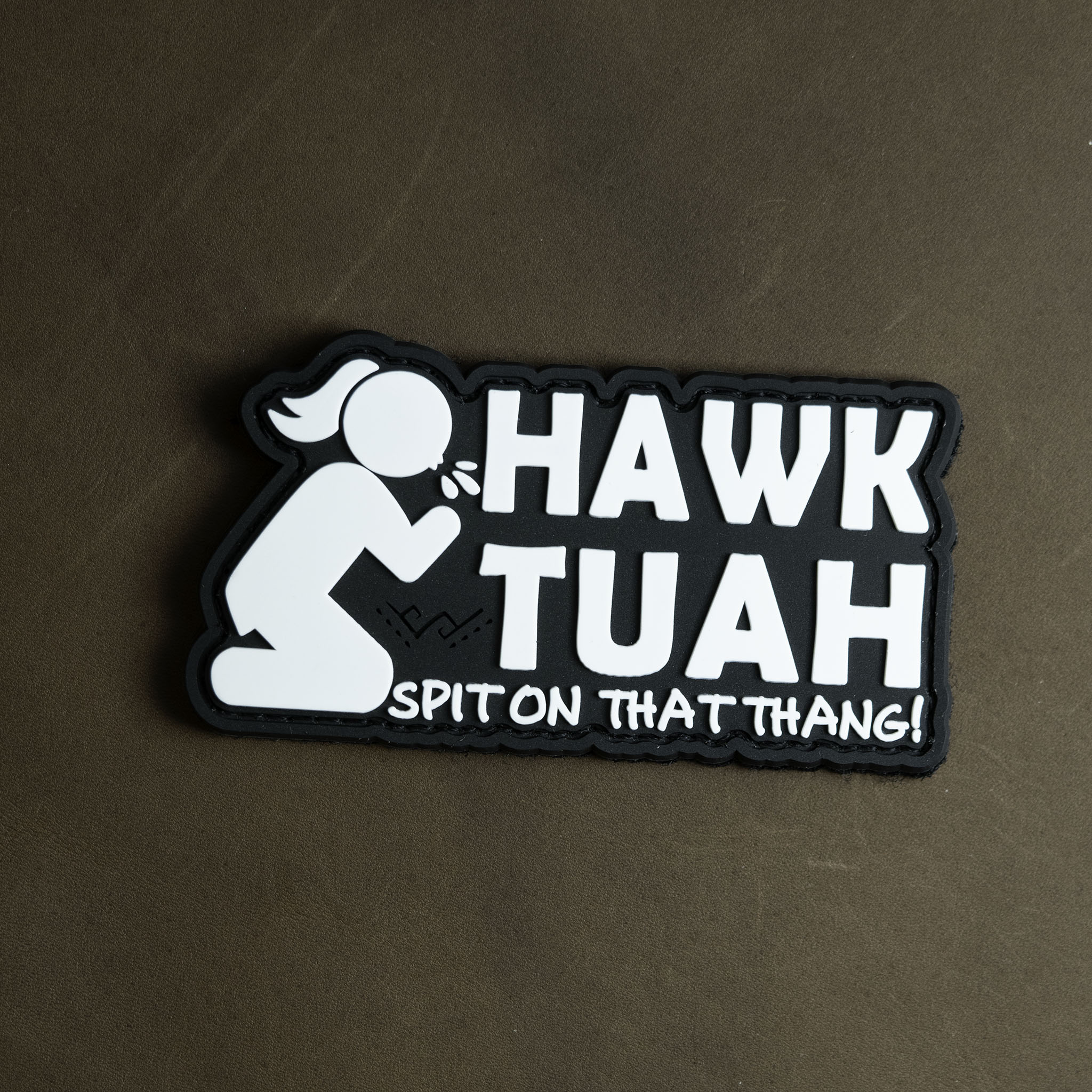 Black and white PVC patch featuring the phrase “Hawk Tuah – Spit On That Thang!” with a kneeling cartoon figure.