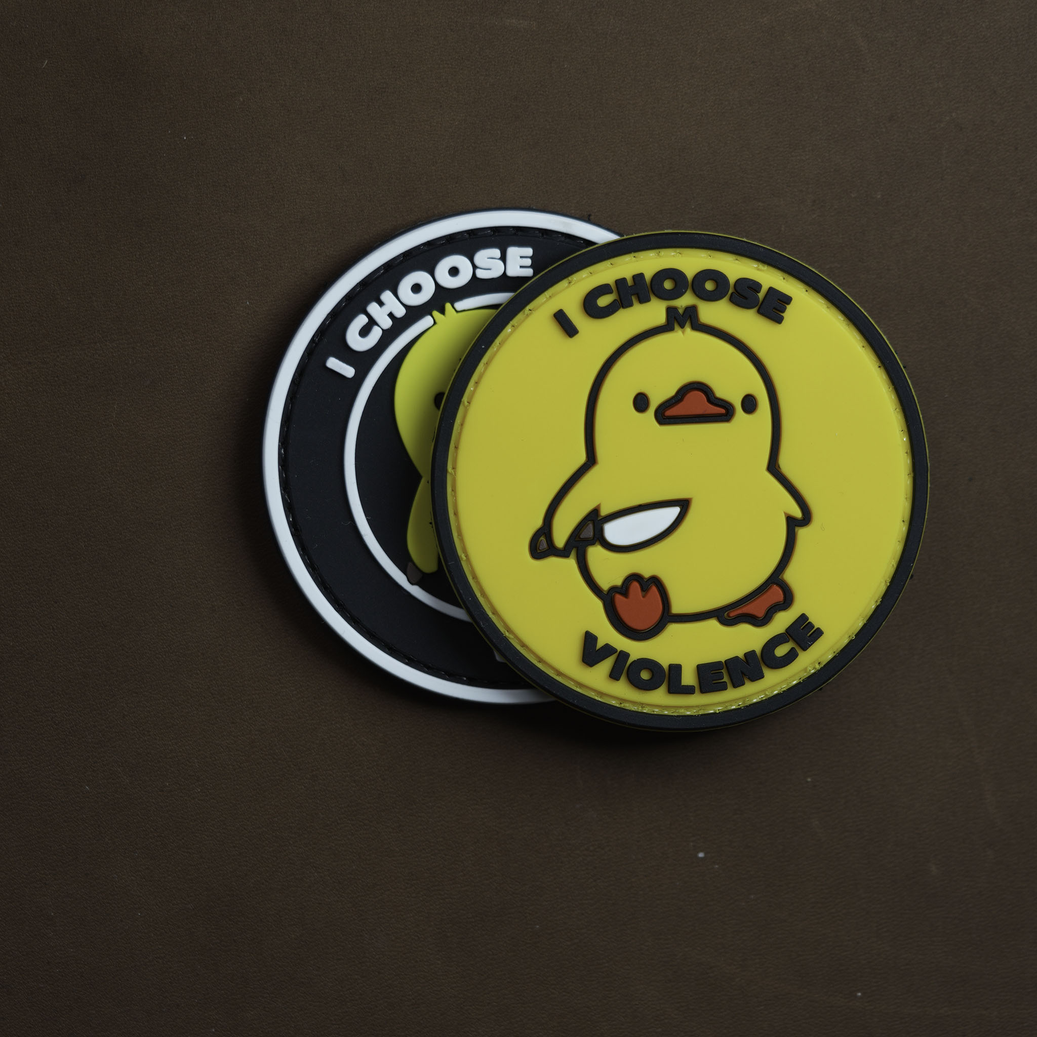 Two round PVC patches featuring a yellow duck holding a knife with the text ‘I CHOOSE VIOLENCE,’ one in yellow and one in black, placed on a brown surface.