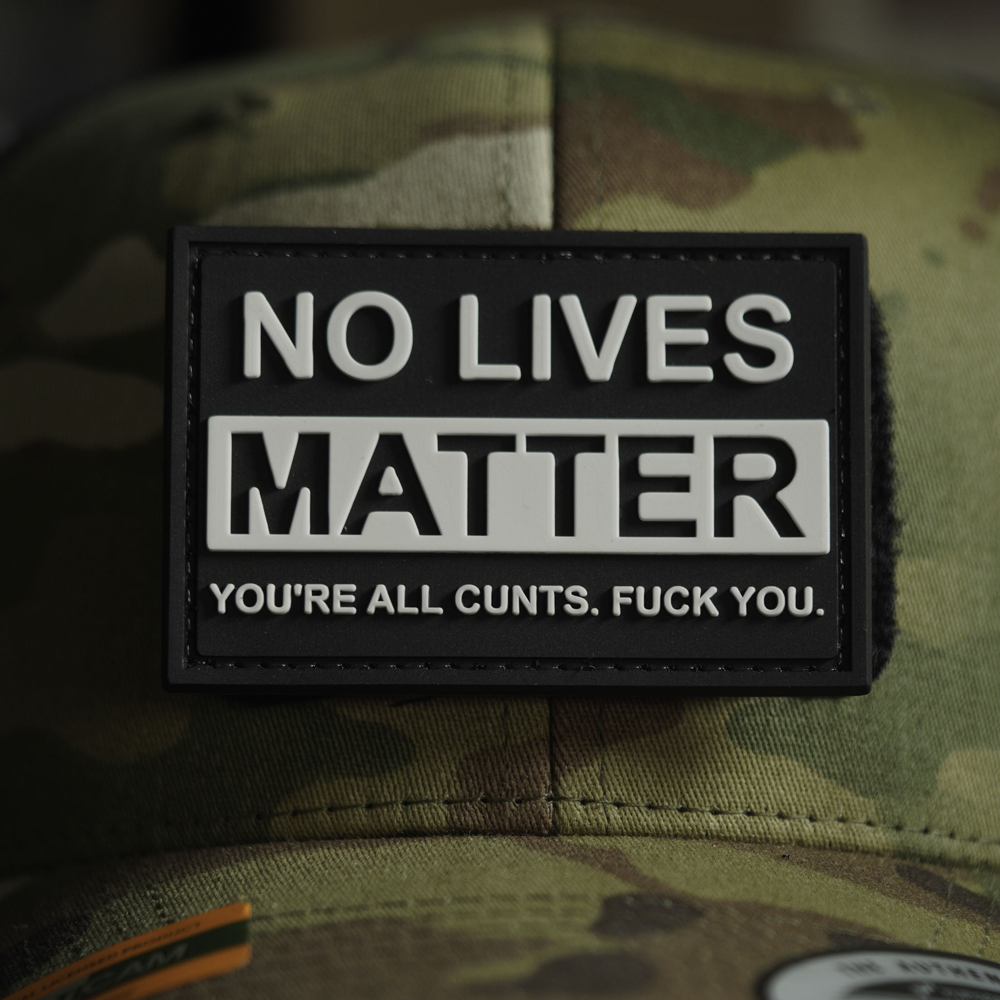 Black PVC patch with raised white text reading ‘NO LIVES MATTER’ and an additional offensive line of text, attached to a camouflage cap.