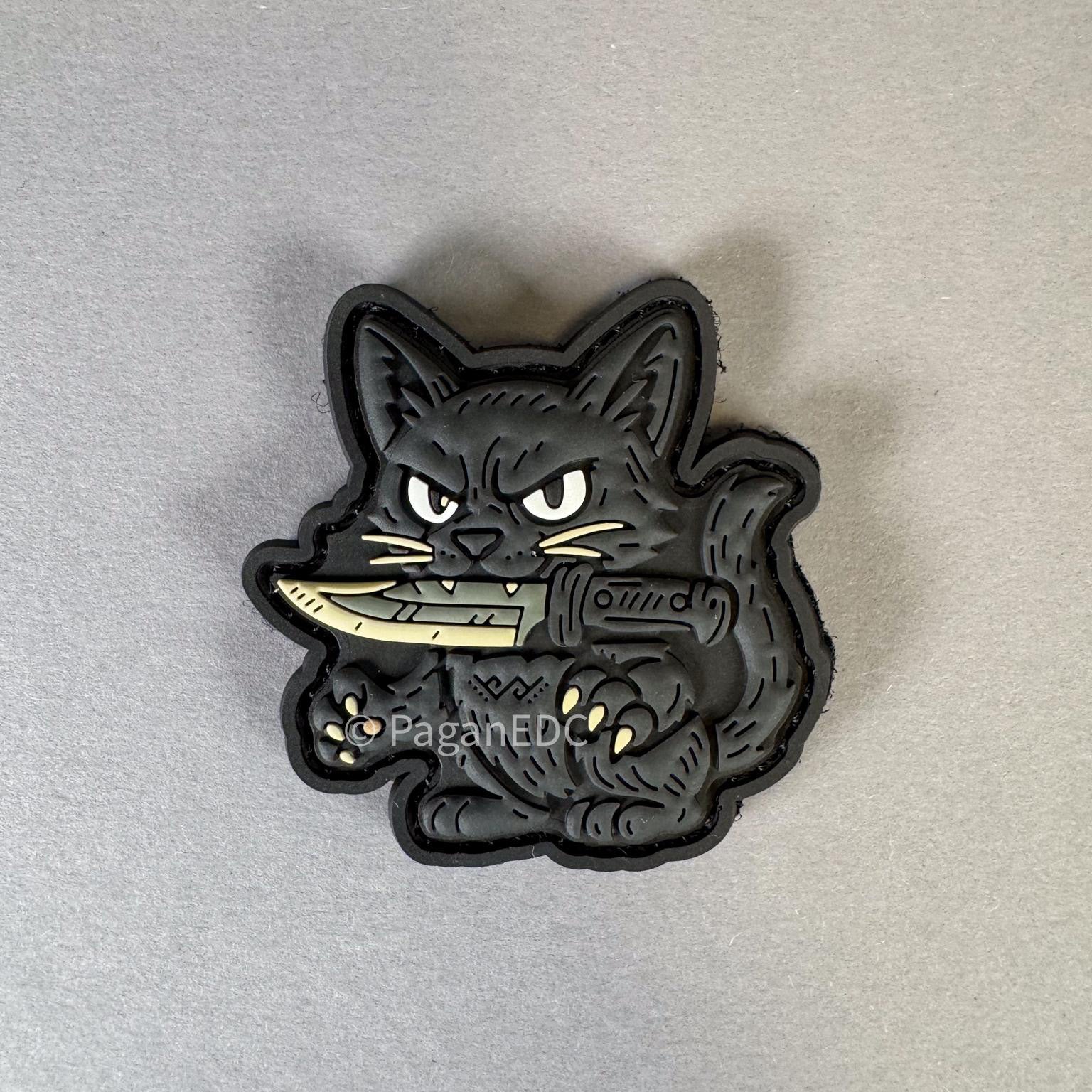 PVC patch featuring a black cat holding a knife, designed by PaganEDC, with glow-in-the-dark eyes and 3D detailing on a gray background.