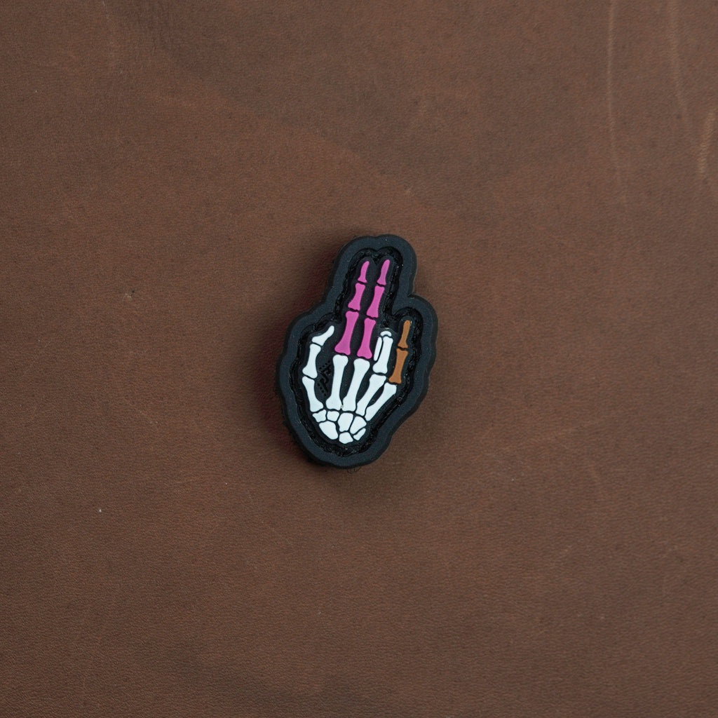 Skeletal hand PVC patch in color, featuring white bones with purple finger segments and a small gold detail, displayed on a brown background