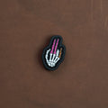 Skeletal hand PVC patch in color, featuring white bones with purple finger segments and a small gold detail, displayed on a brown background