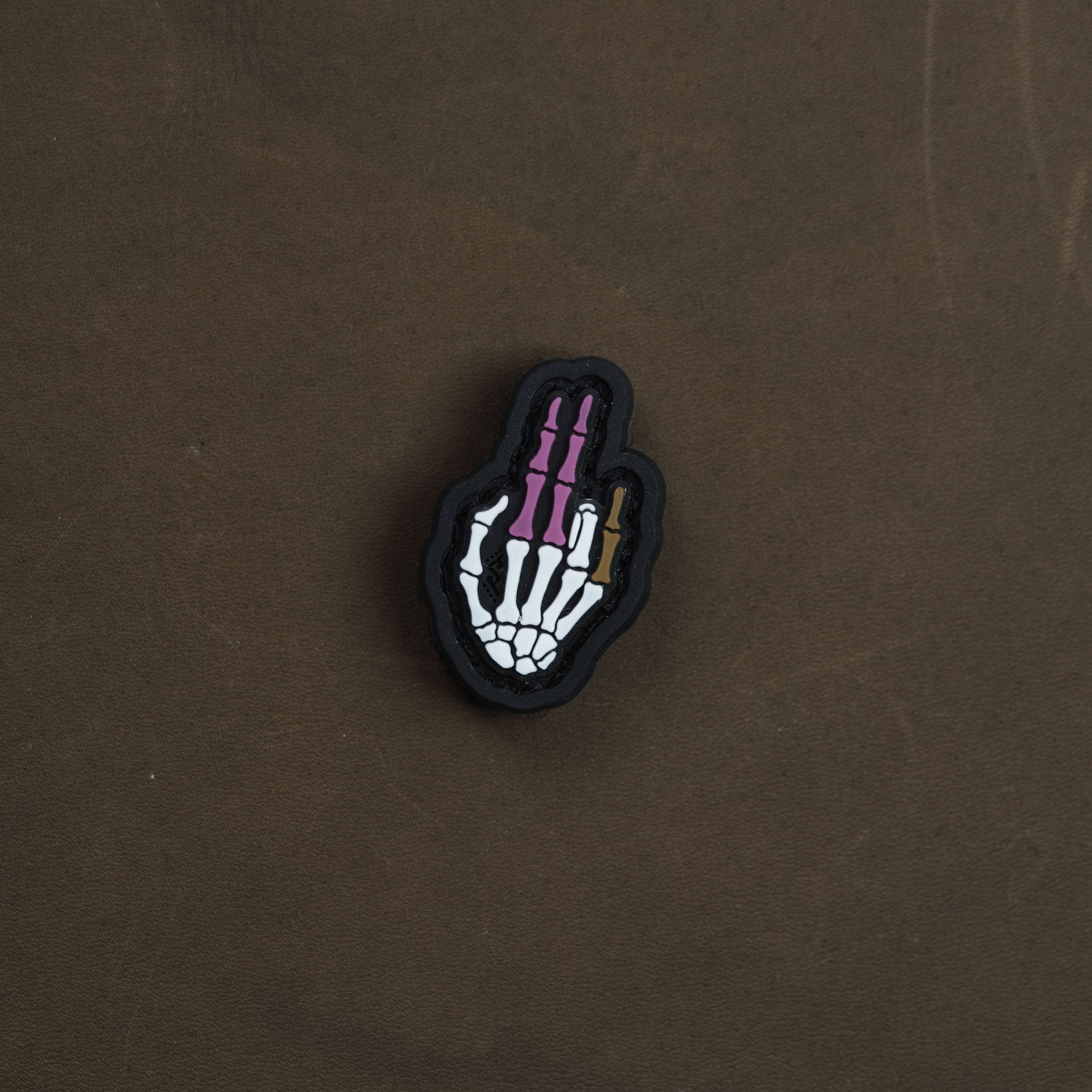 Skeletal hand PVC patch in color, featuring white bones with purple finger segments and a small gold detail, displayed on a brown background