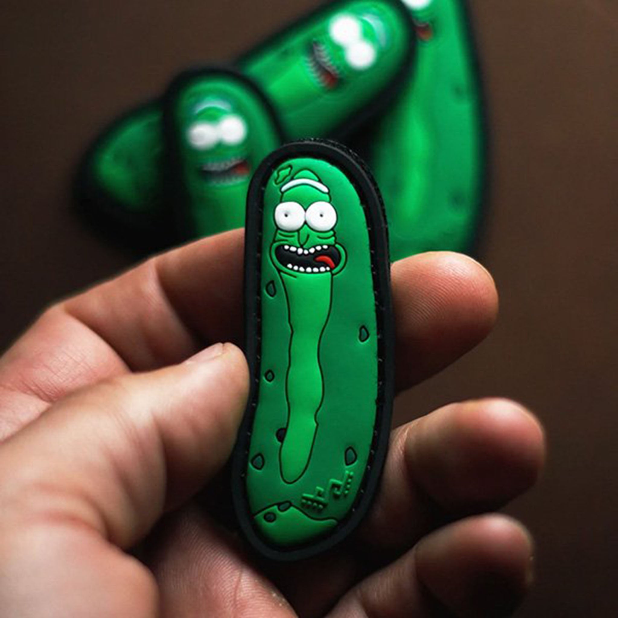 Green pickle-shaped PVC patch with a cartoon face, held between fingers, with additional identical patches blurred in the background
