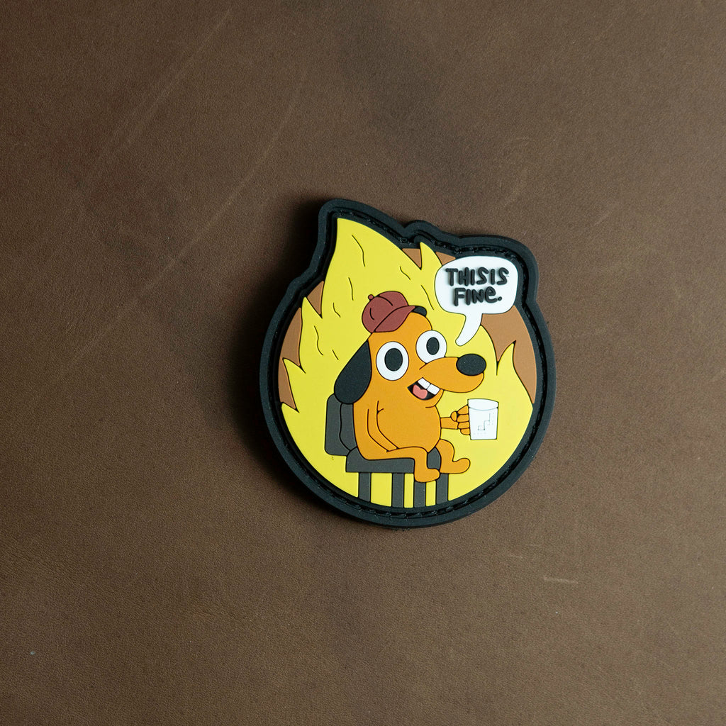 PVC patch depicting the ‘This Is Fine’ dog sitting calmly in a burning room with a speech bubble saying ‘This is fine,’ shown on a brown background