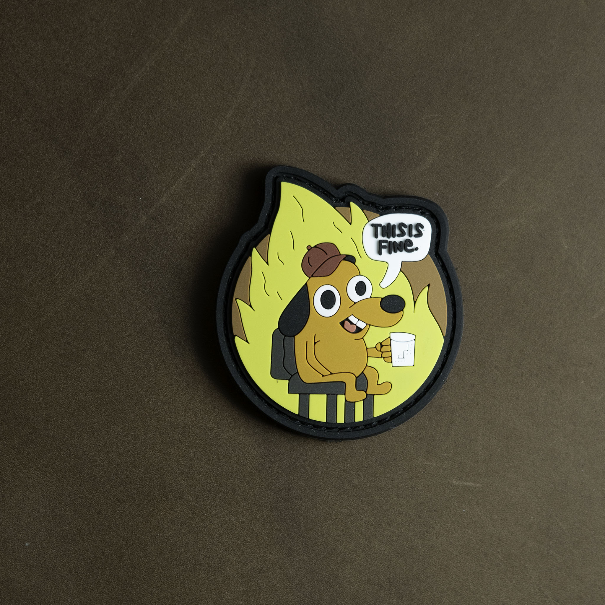 PVC patch depicting the ‘This Is Fine’ dog sitting calmly in a burning room with a speech bubble saying ‘This is fine,’ shown on a brown background