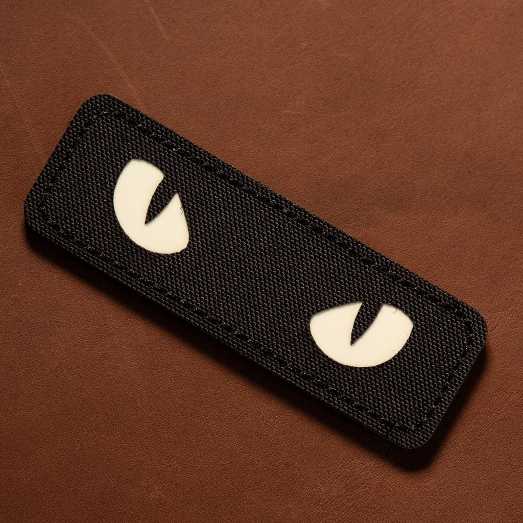 Black woven patch featuring two white reflective cat eyes on a brown background.
