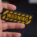 Hand holding a rectangular black reflective patch with yellow honeycomb-pattern caution stripes and the word “WARNING” in bold reflective lettering.