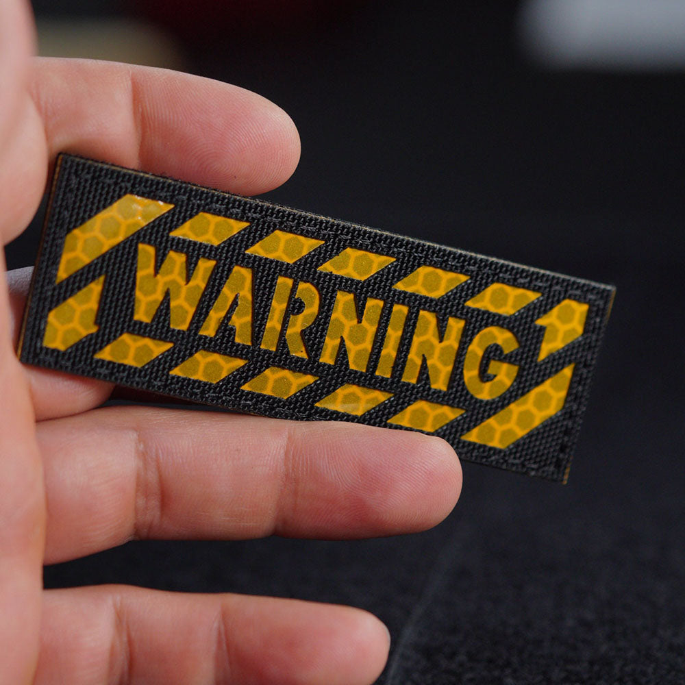 Hand holding a rectangular black reflective patch with yellow honeycomb-pattern caution stripes and the word “WARNING” in bold reflective lettering.